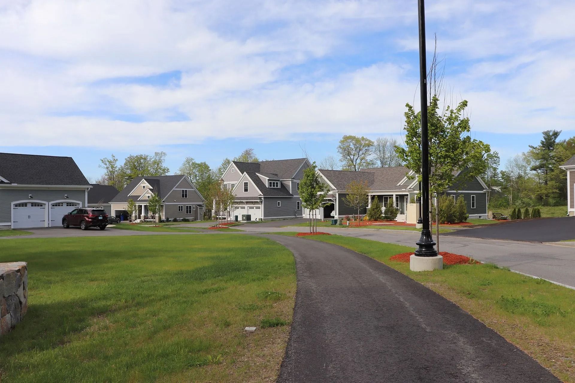 Paved driveway leading to several gray houses with manicured lawns under a partly cloudy sky.