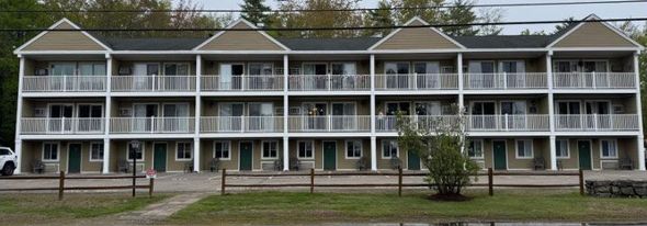 Three-story tan motel building with white balconies and green doors, in front of a wooden fence and a grassy area.