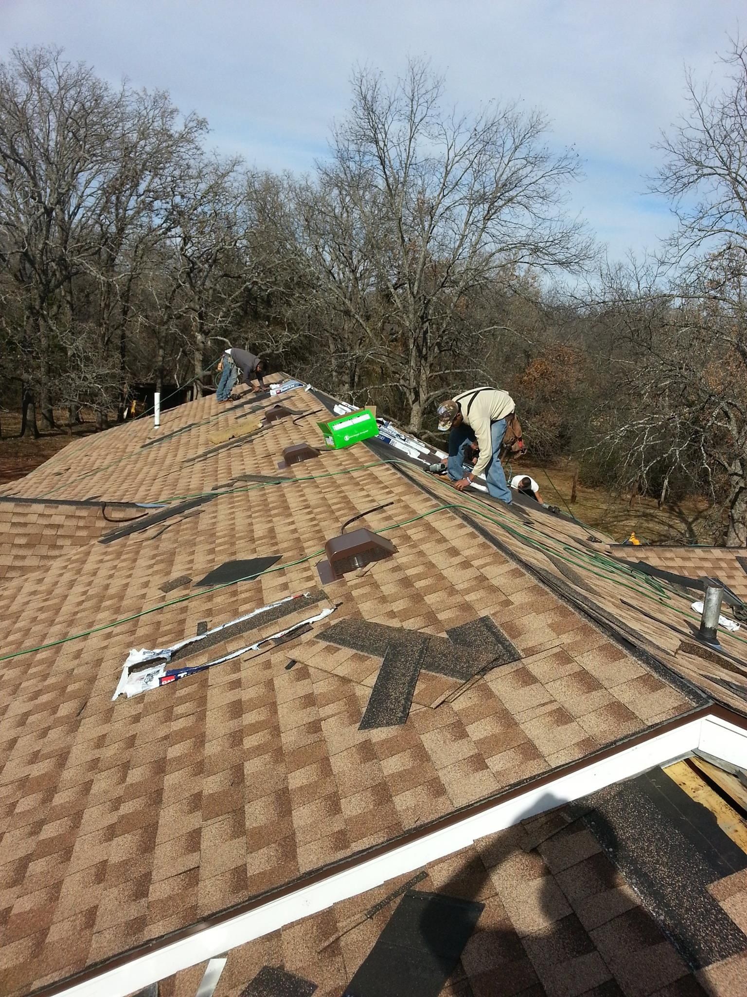 Roofers installing solar panels on a brown shingled roof with trees in the background on a sunny day.