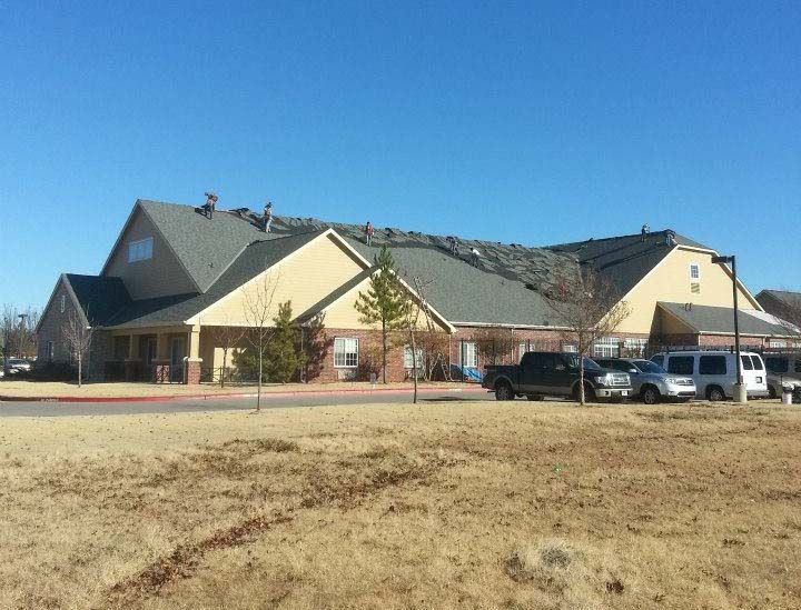 Building with damaged roof; brown grass, blue sky, several vehicles in parking area.