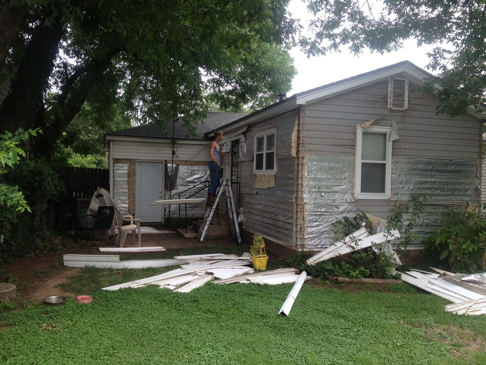 Person on ladder working on the exterior of a small house with siding removed, surrounded by debris.