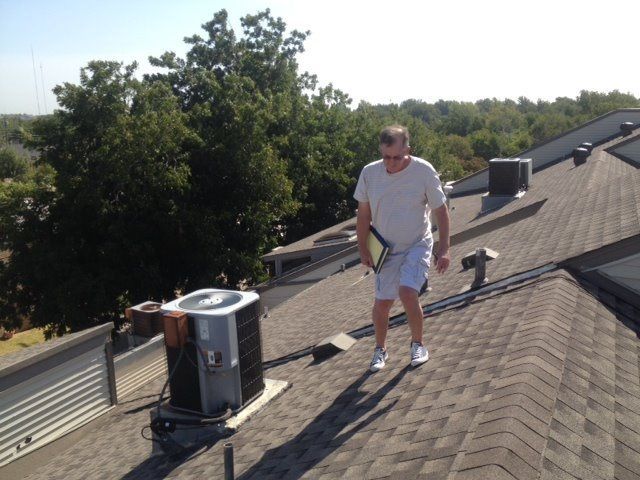 Man in shorts and white shirt walking on a roof, near an AC unit. Sunny day, trees in the background.