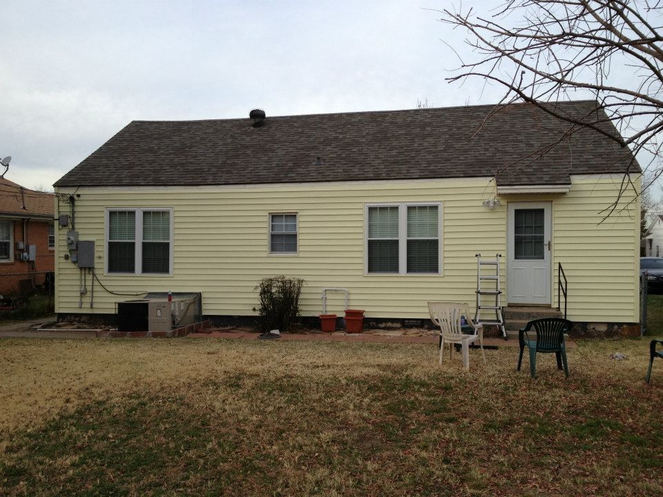 Back of a small yellow house with brown roof, windows, and a door, in a yard with dry grass.