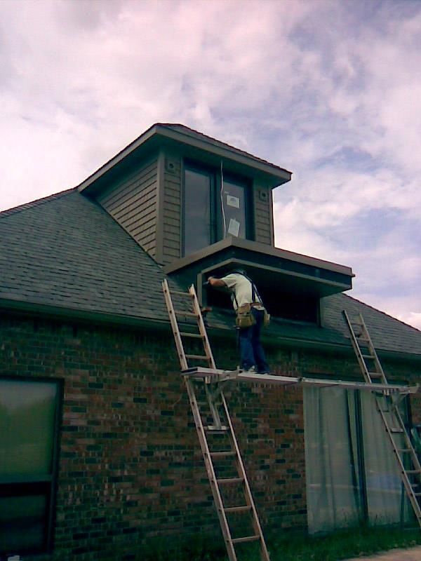 Person on scaffolding installing window in brown-sided dormer on brick house; cloudy sky.