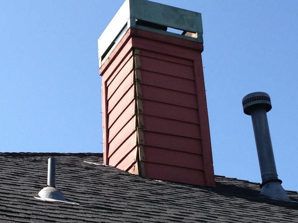 Red chimney with a copper cap on a black shingled roof against a clear blue sky.