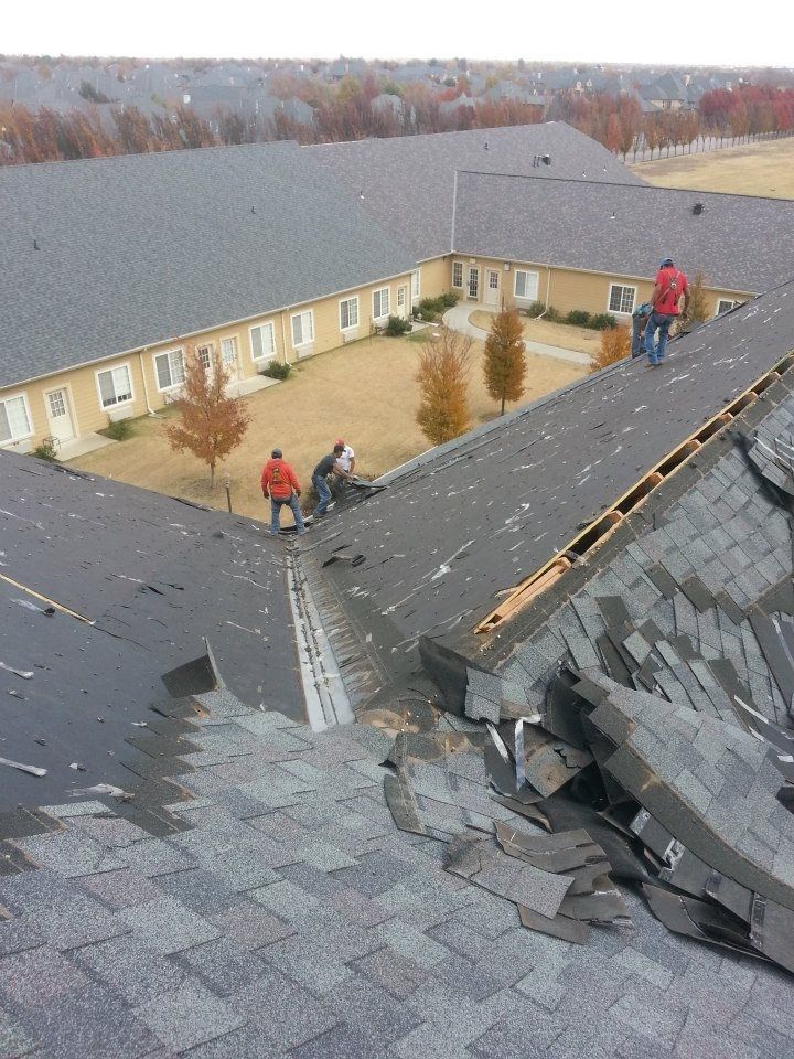 Roofers repairing a damaged shingle roof on a building with a courtyard, trees, and gray shingles.