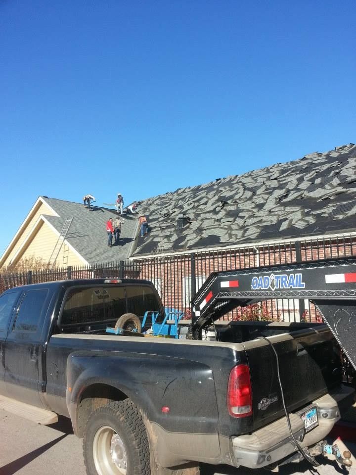 Workers on a roof removing old shingles next to a pickup truck and trailer under a blue sky.