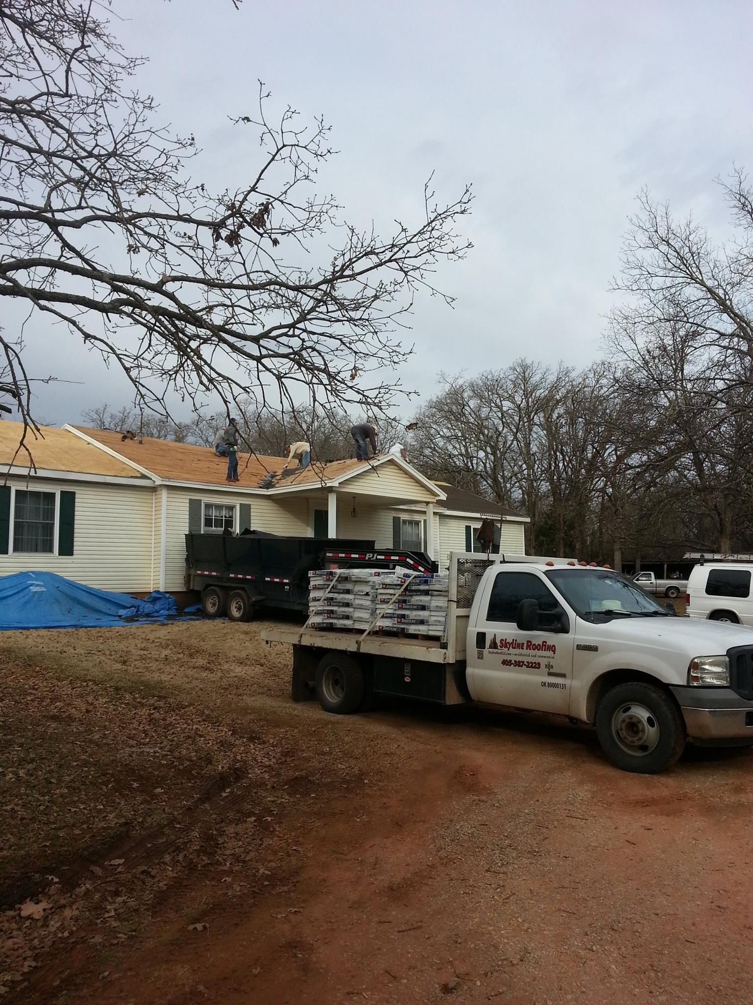 A flatbed truck parked in front of a house, with roofing materials visible and a roof partially uncovered.