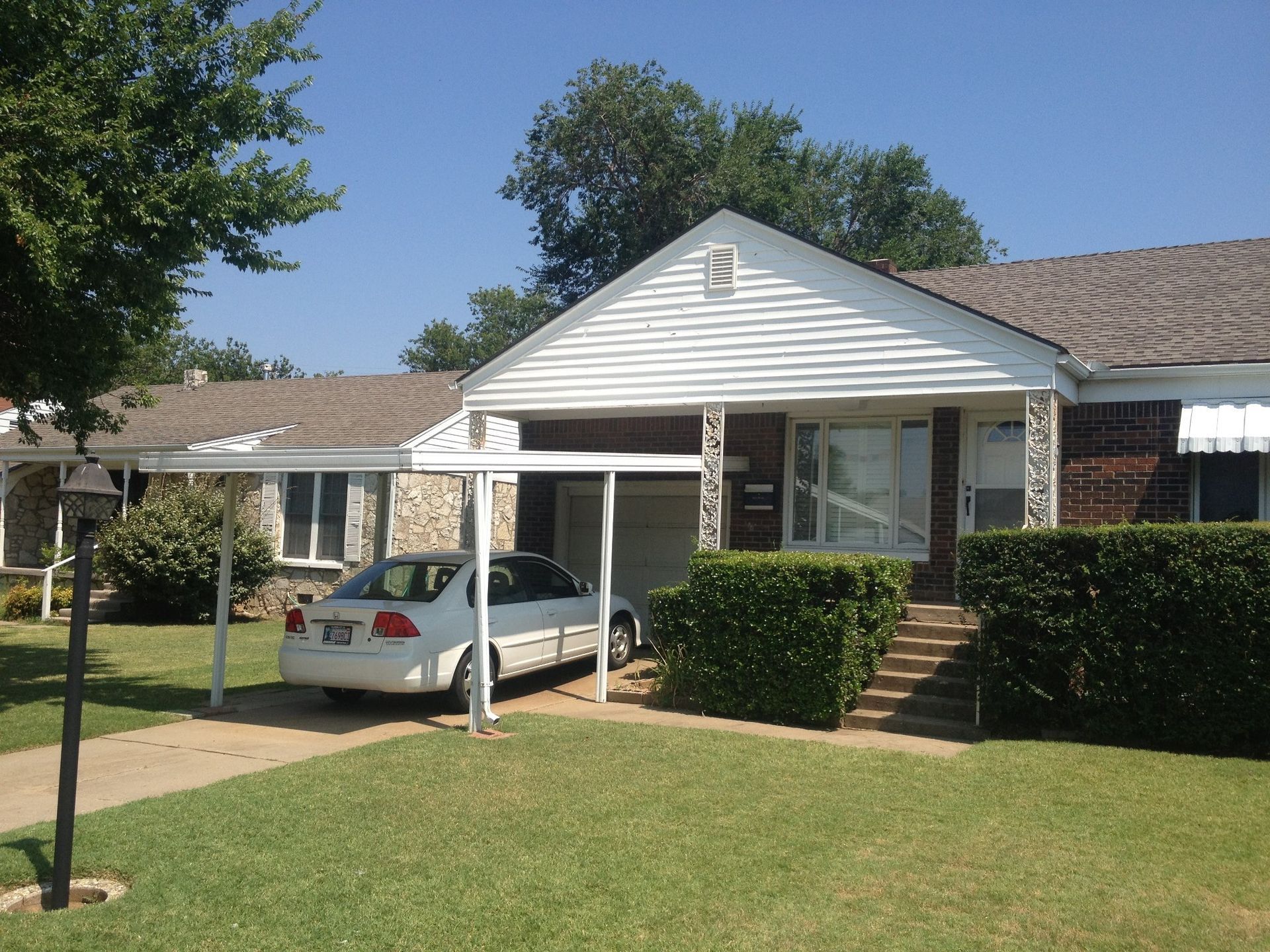 White car parked under a carport of a single-story house with green lawn and blue sky.