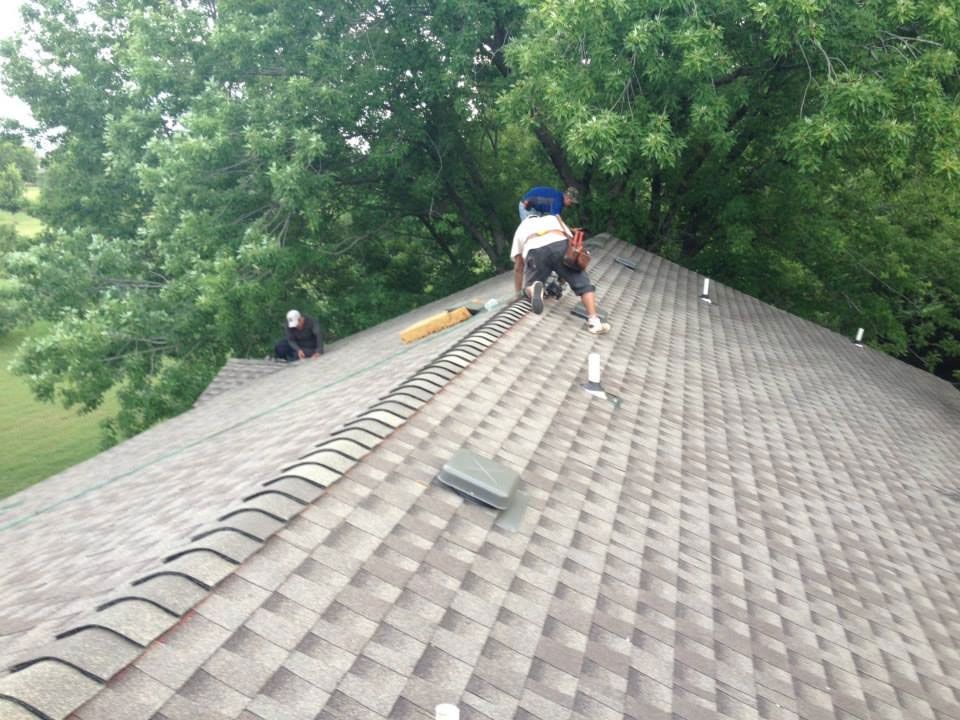 Roofers installing shingles on a house roof, with tools and a tree in the background.