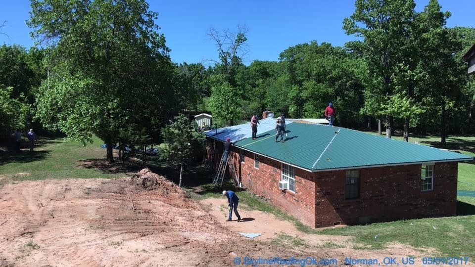 Workers replacing a green metal roof on a brick house surrounded by trees.