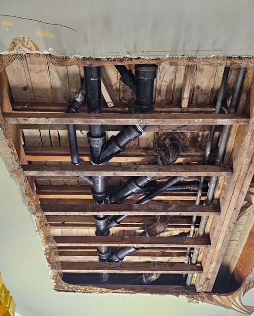 Exposed plumbing pipes of various sizes and fittings, seen from below, in a wood-framed ceiling.