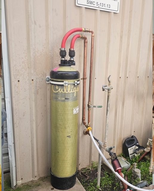 Water filtration system against a beige wall; copper pipes and red hoses connect to the filter.
