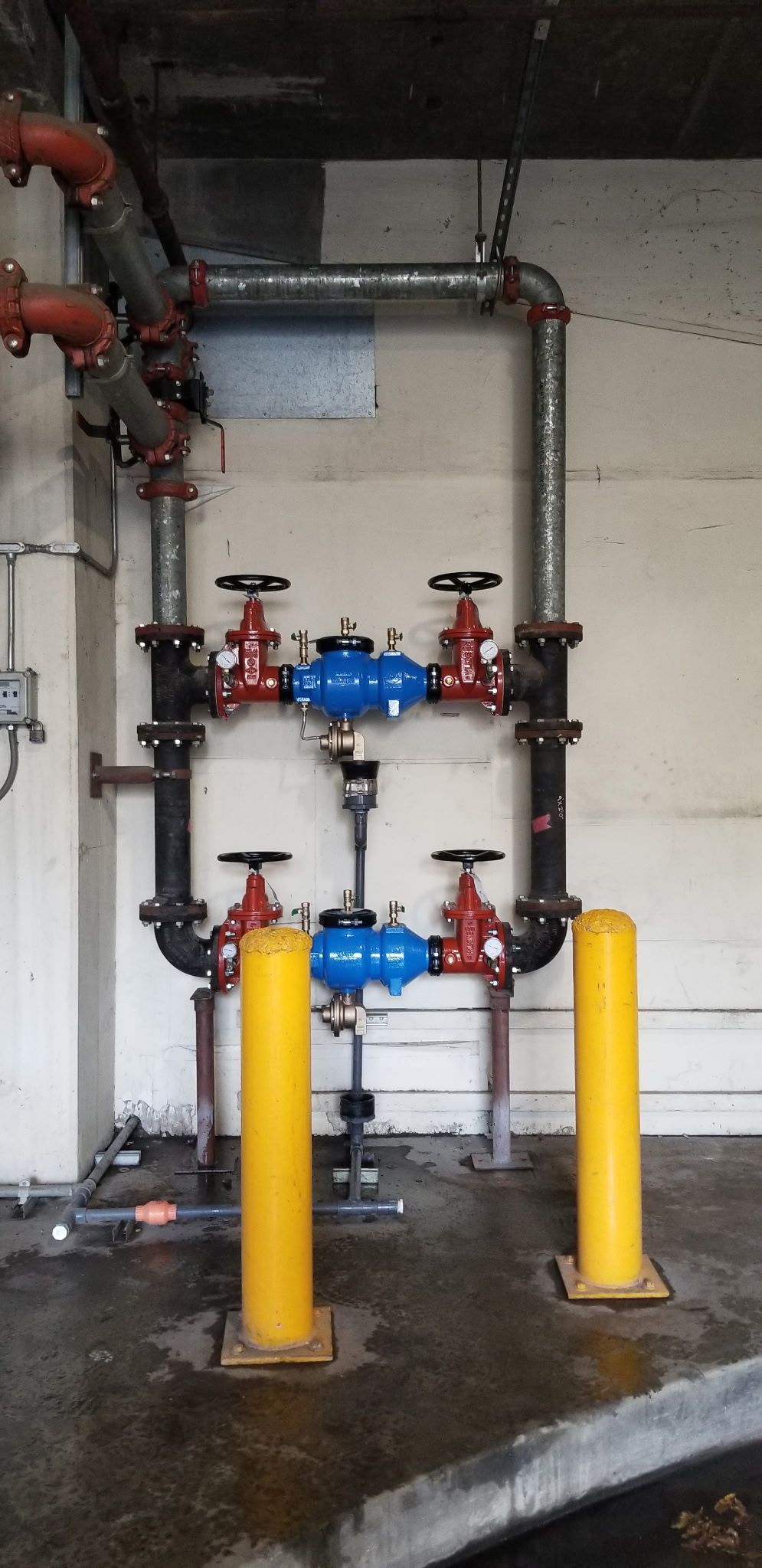 Pipes with valves and blue water meters, protected by yellow bollards, on a wet floor.