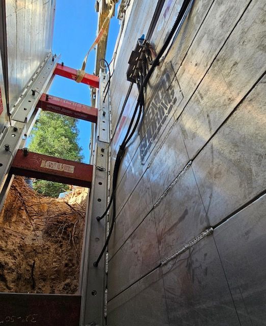 Trench shoring with metal panels, ladders, and utilities within the excavated space, angled upward towards a blue sky.