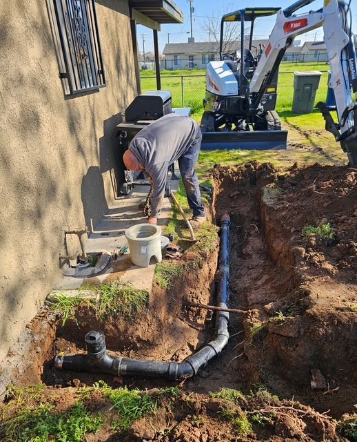 Man working on underground plumbing pipes in a trench near a house, with an excavator in the background.