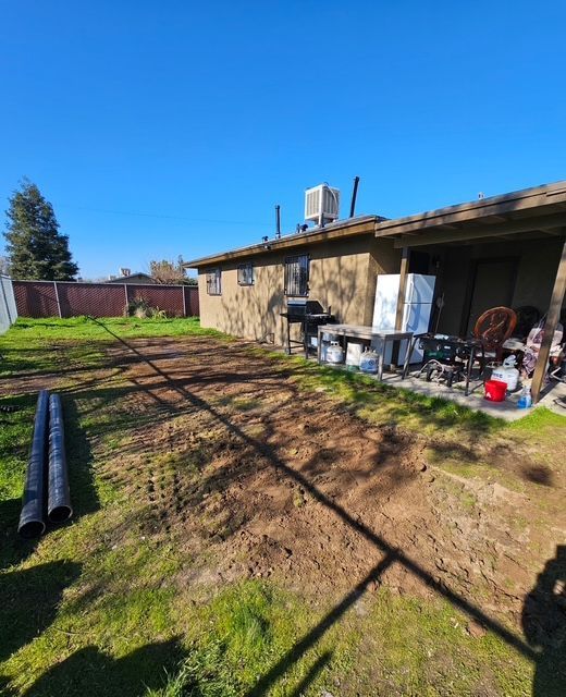Backyard with a house, cleared dirt area, grass, pipes, and a clear blue sky.