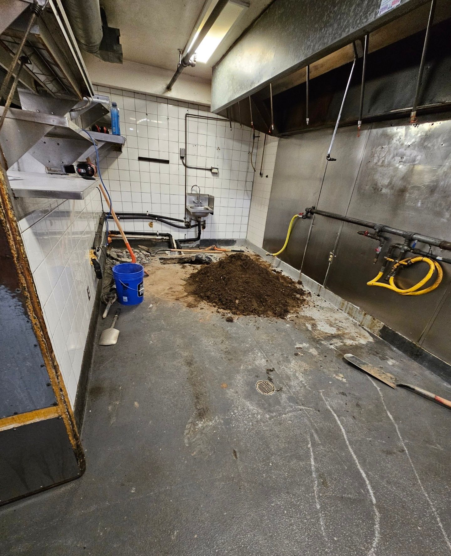 A cluttered room with a pile of dirt. The floor is gray. Includes equipment, pipes, and a bucket.