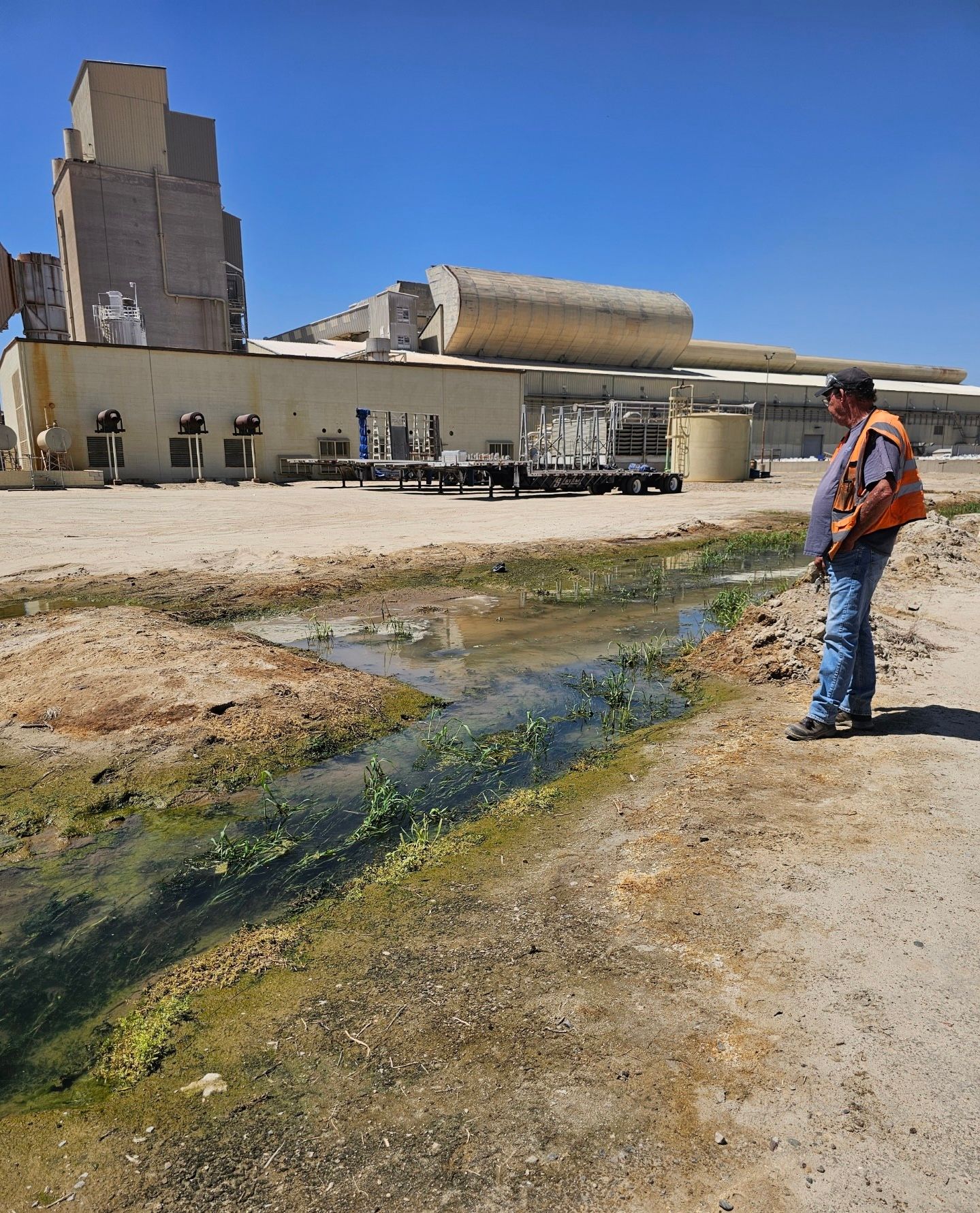Man in vest looks at polluted waterway near a dilapidated industrial building under a blue sky.