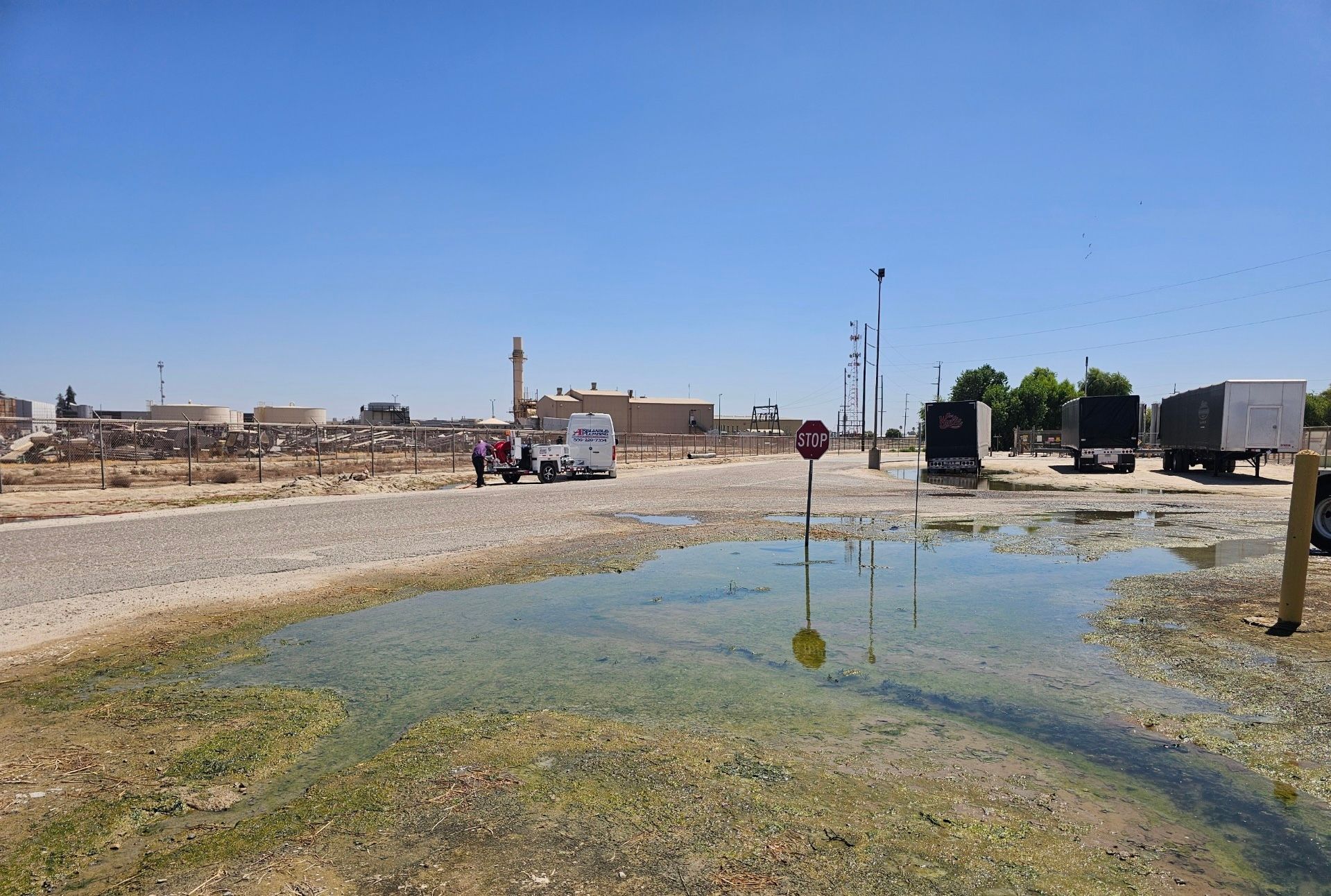 Puddle on gravel road, with parked trucks, stop sign, and distant buildings under blue sky.