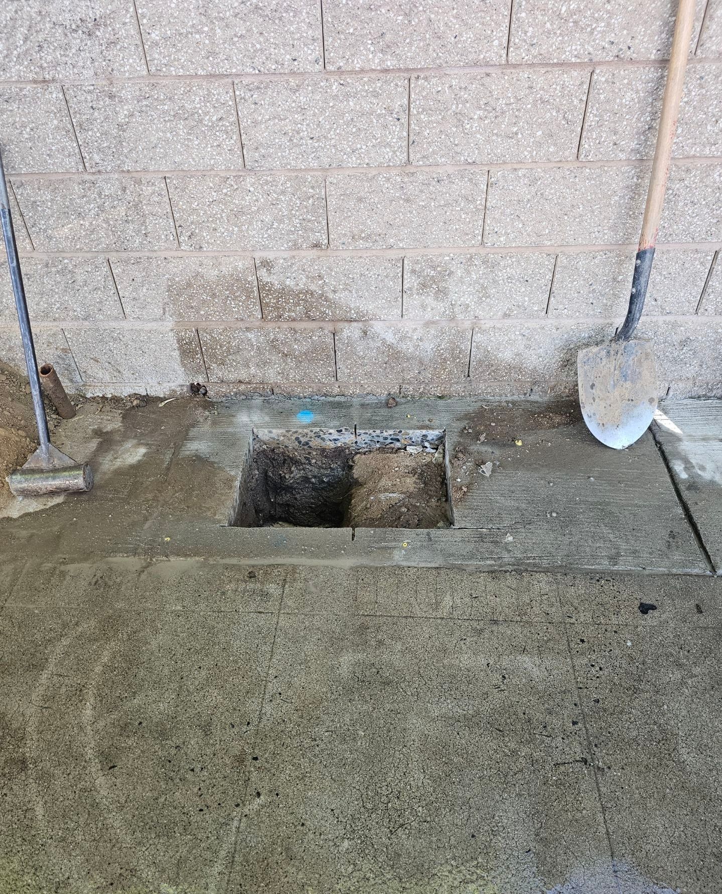 Square hole cut in concrete near a cinder block wall, with tools resting nearby.