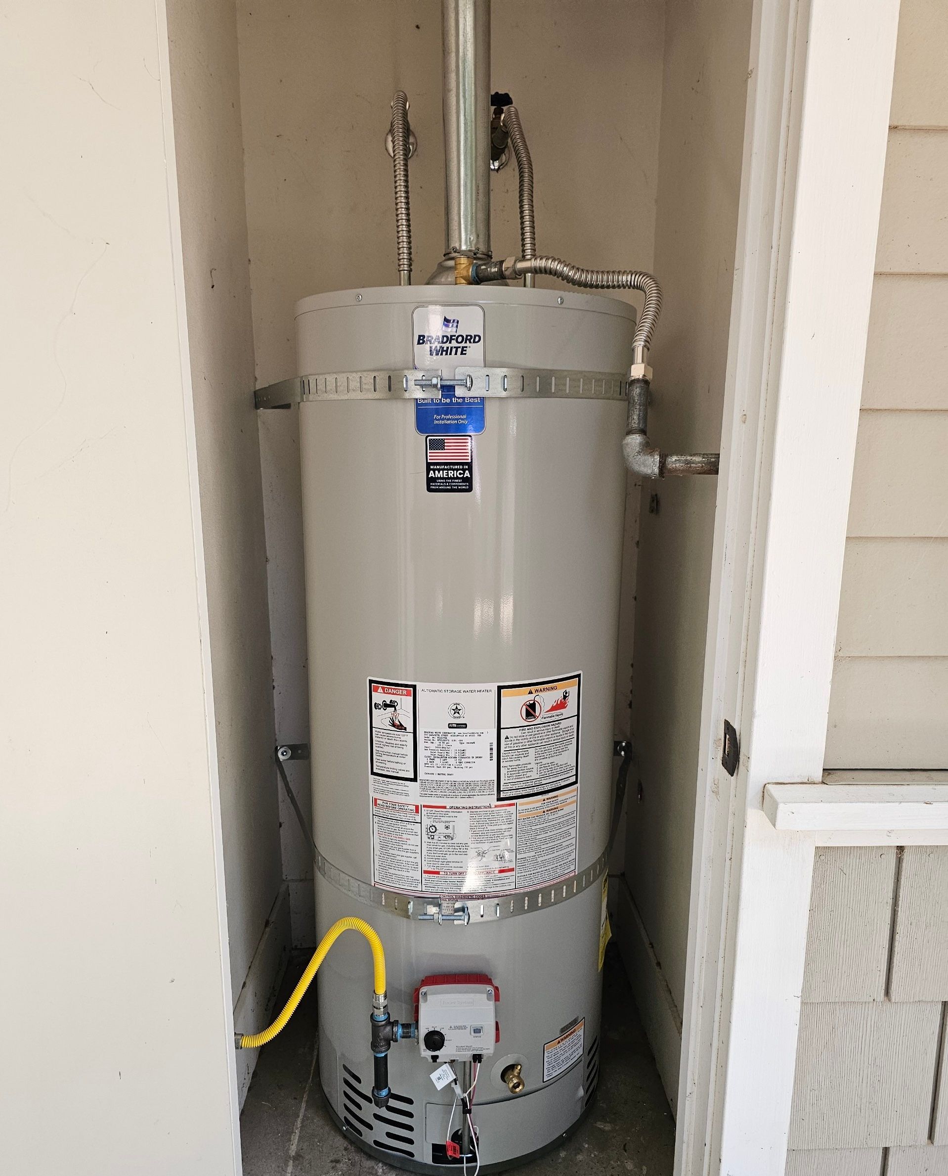 A water heater in a white recess, with silver tank, pipes, and yellow gas line.