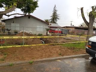 A fenced yard with a dirt pit marked with caution tape. A house and cars are in the background.