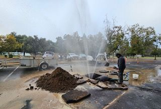 Water main break, person standing, water spraying, asphalt, dirt pile, cars in background.
