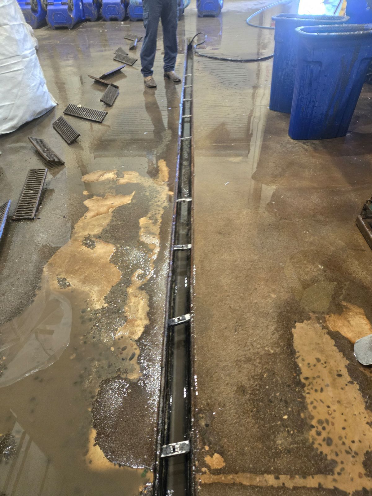 A person stands over a long, open drain in a wet industrial floor. Various objects lie nearby.
