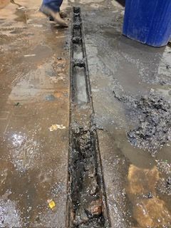 Dirty floor drain in a concrete floor. Mud and debris around the drain. A person's feet are visible in the background.