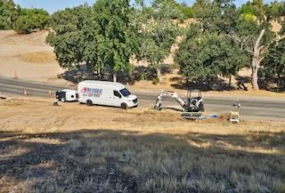 A white utility van with a trailer and small excavator working near a road.