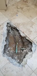 Hole in tiled floor reveals plumbing. Broken tiles, concrete, and copper pipes are visible.