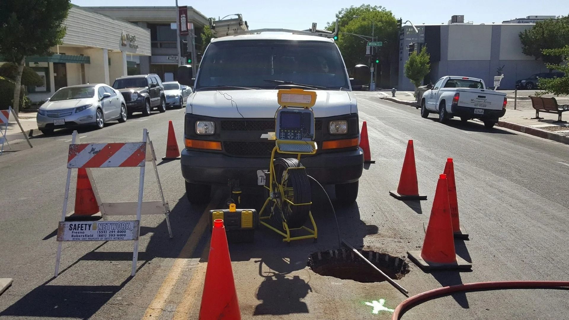 Van parked on street; sewer inspection equipment set up around open manhole; orange cones and street sign are present.