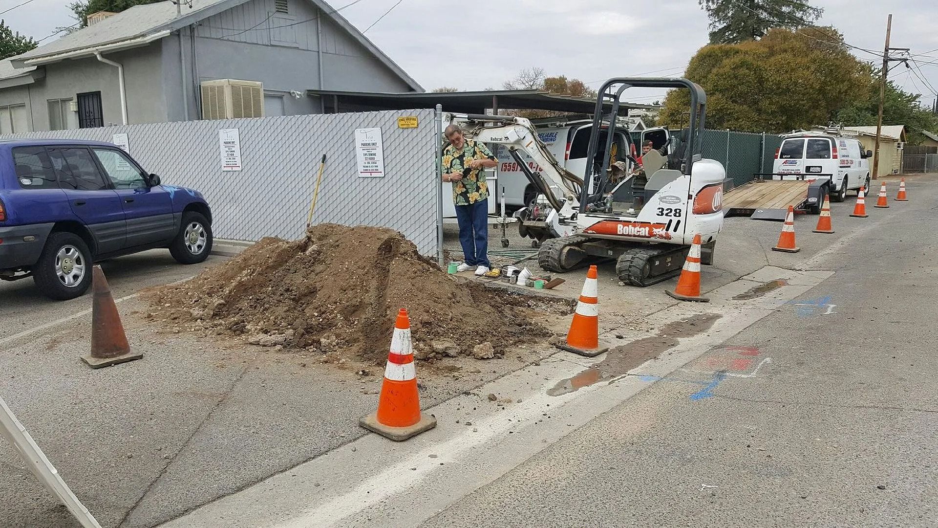 Construction site on a street with an excavator, worker, and orange cones. Blue car parked nearby.