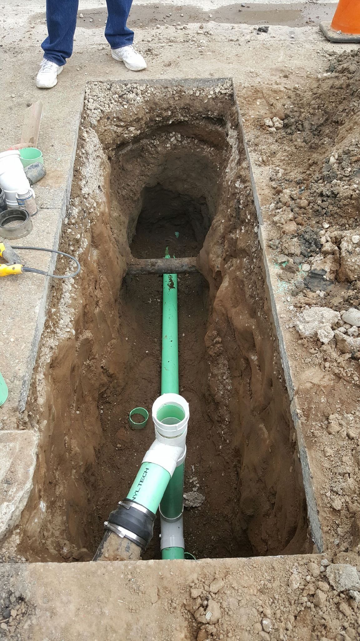 Green pipes in trench, worker's feet visible. Construction site, brown dirt and gravel.