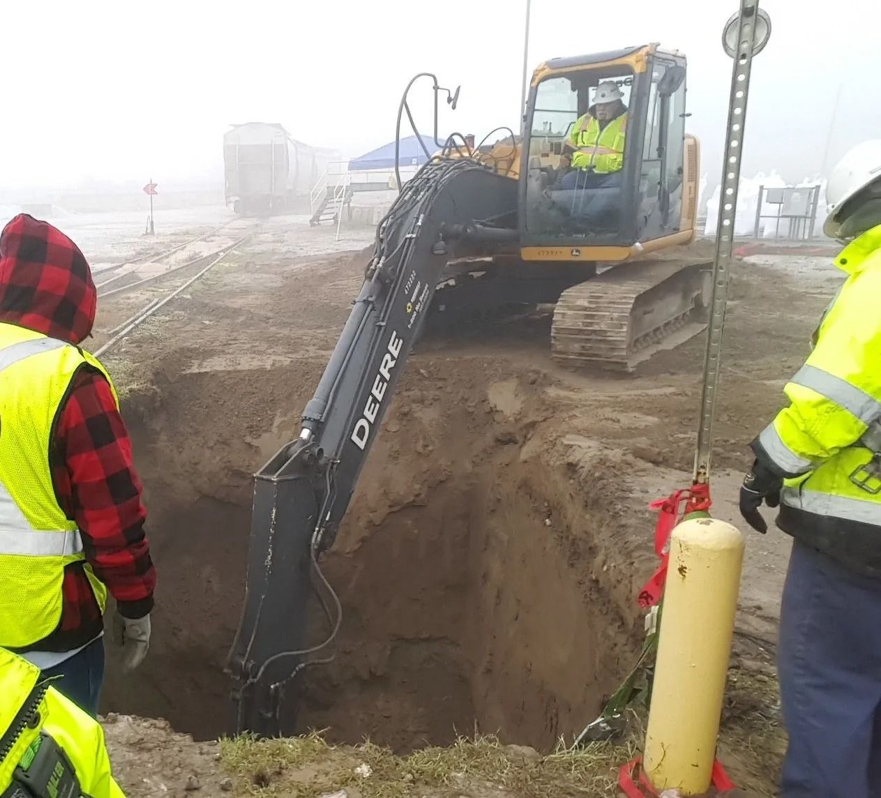 Excavator digging a deep hole, two workers in safety vests observing. Overcast, outdoor setting.