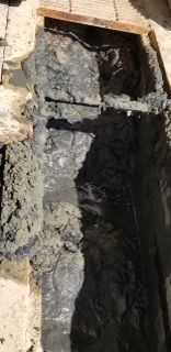 Excavated trench with a dark, possibly buried, pipe. Mud and debris surround it.