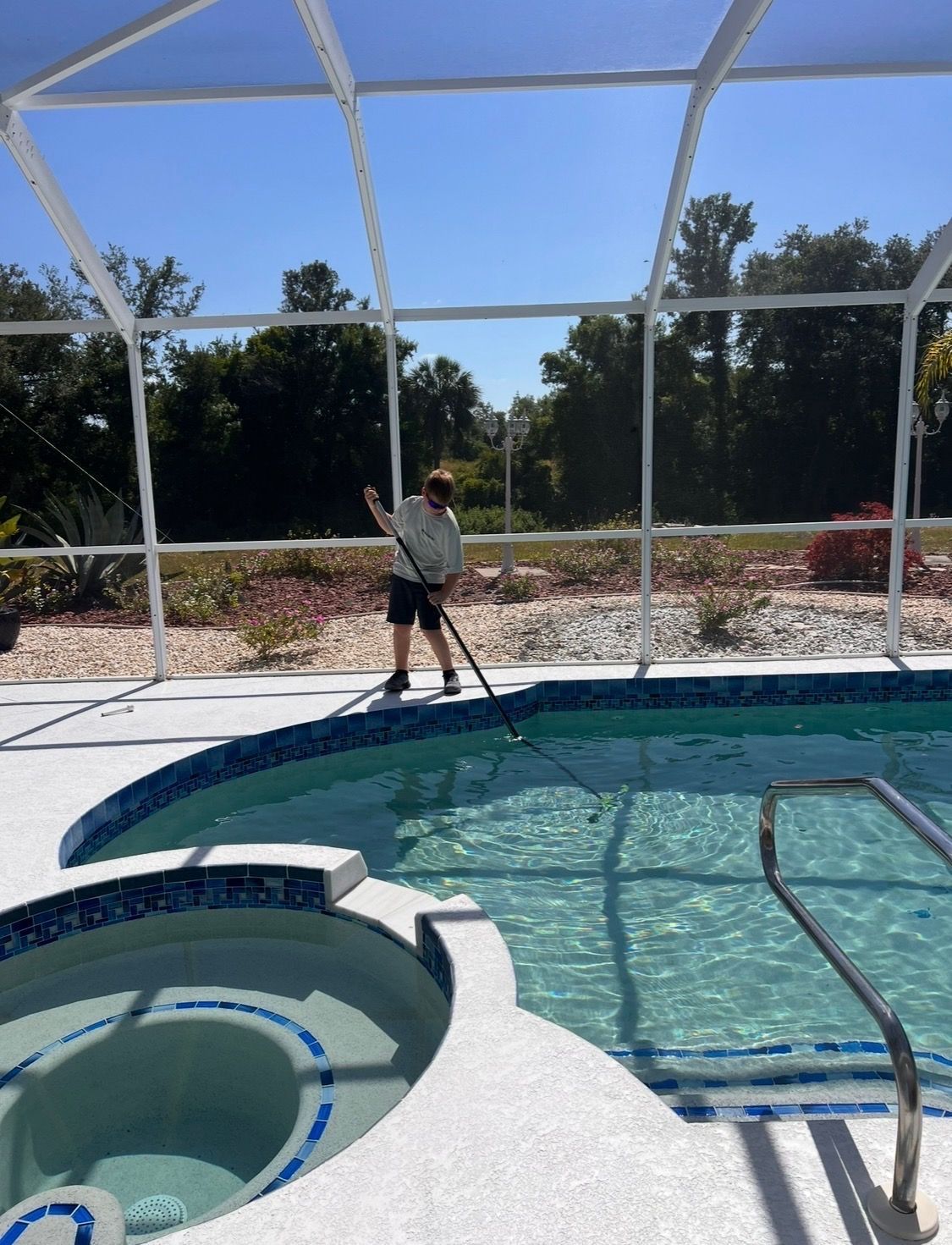 Person standing by a screened-in pool with a vacuum pole, near a hot tub and blue water