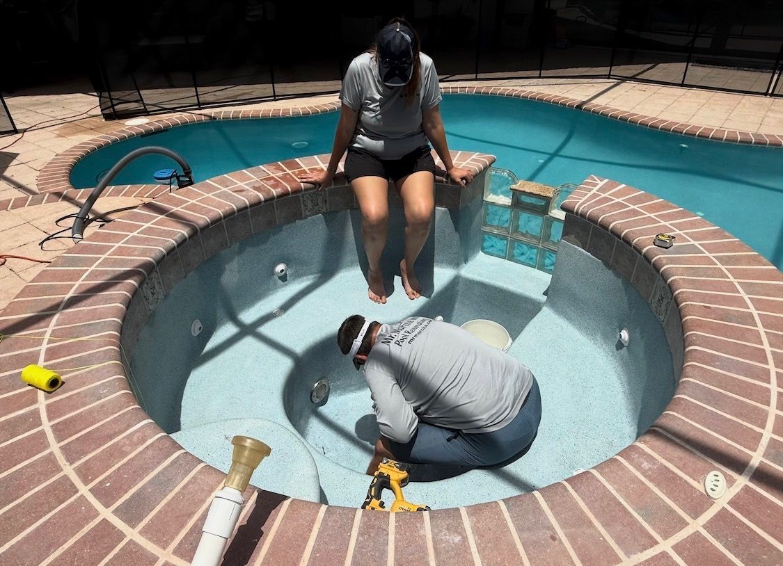 Two people in a small empty pool, one sitting on the edge and one crouched inside, with turquoise water nearby