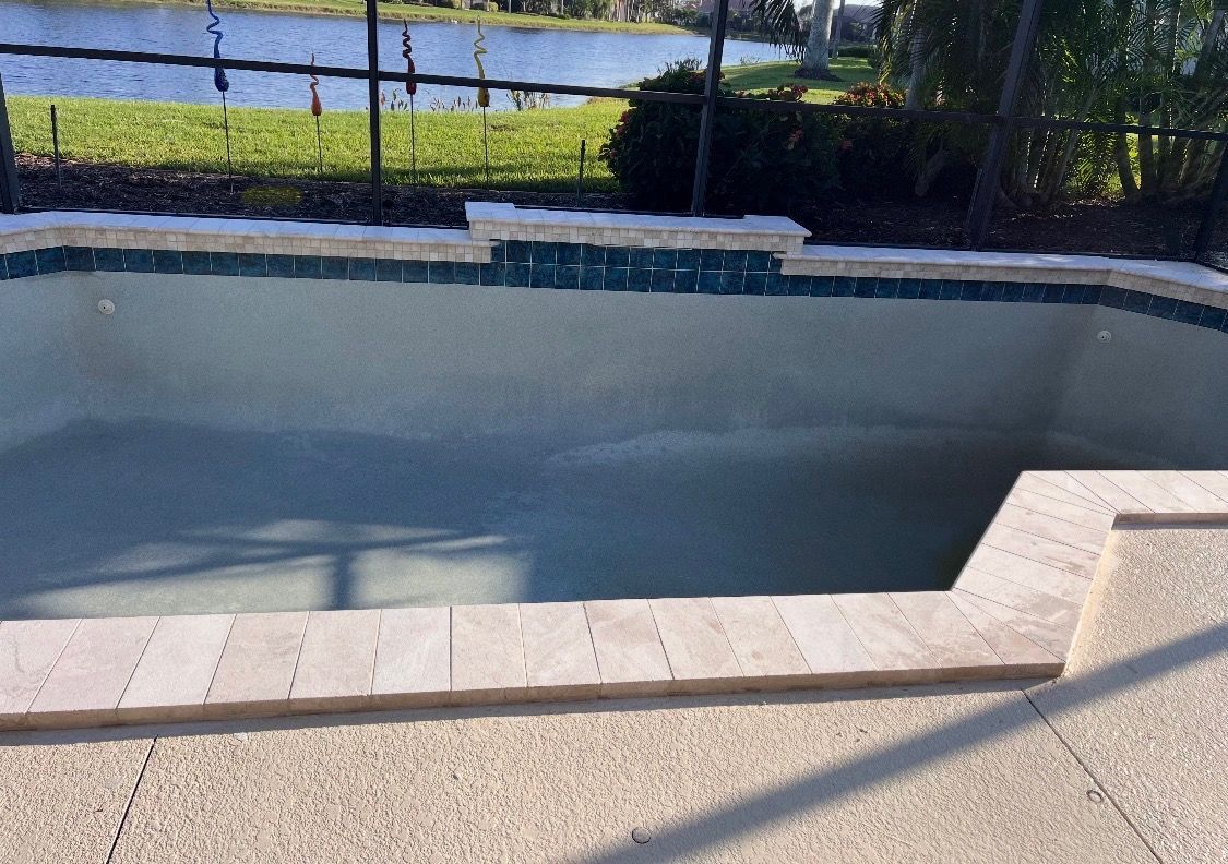 Empty drained pool with concrete steps and sunlit patio beside a fence and water beyond
