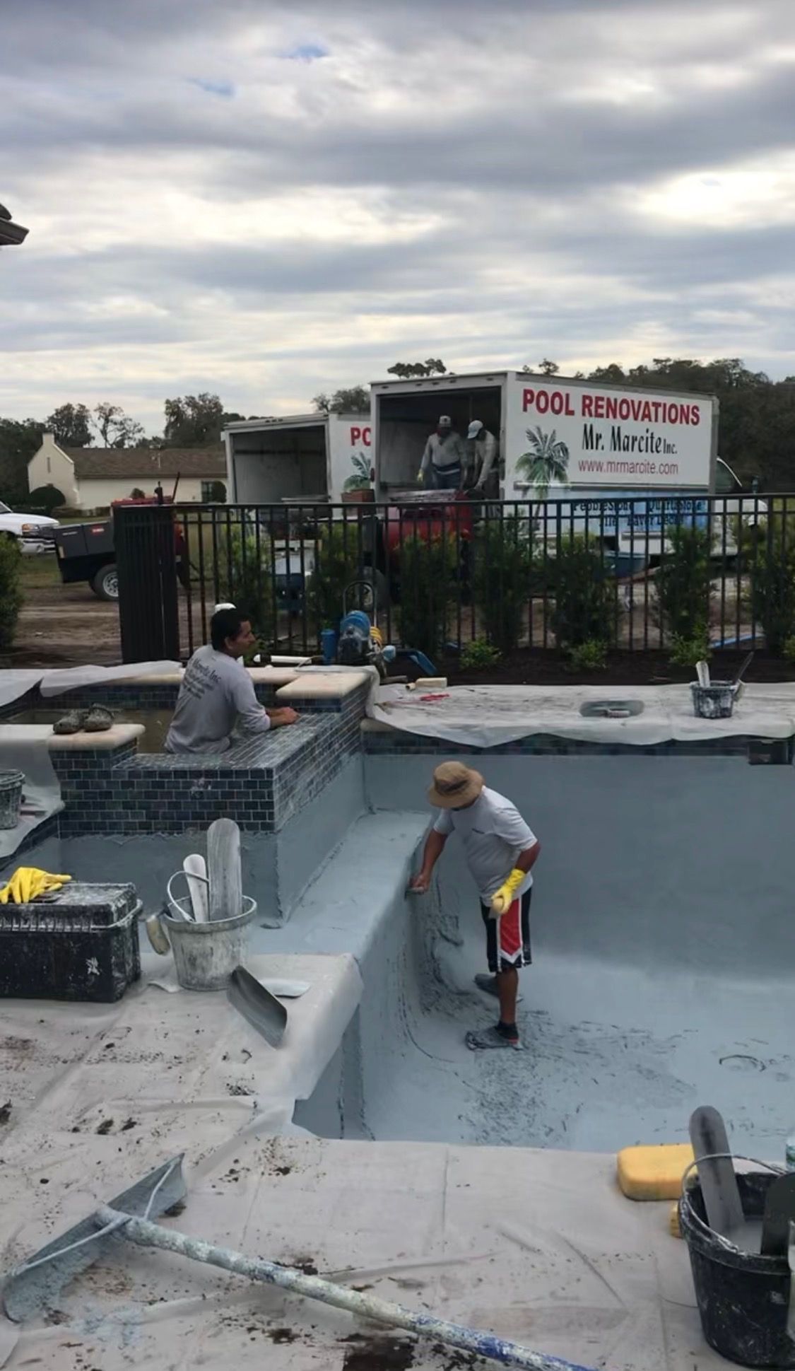 Workers resurfacing a pool deck beside an empty pool, with a fence and cloudy sky in the background