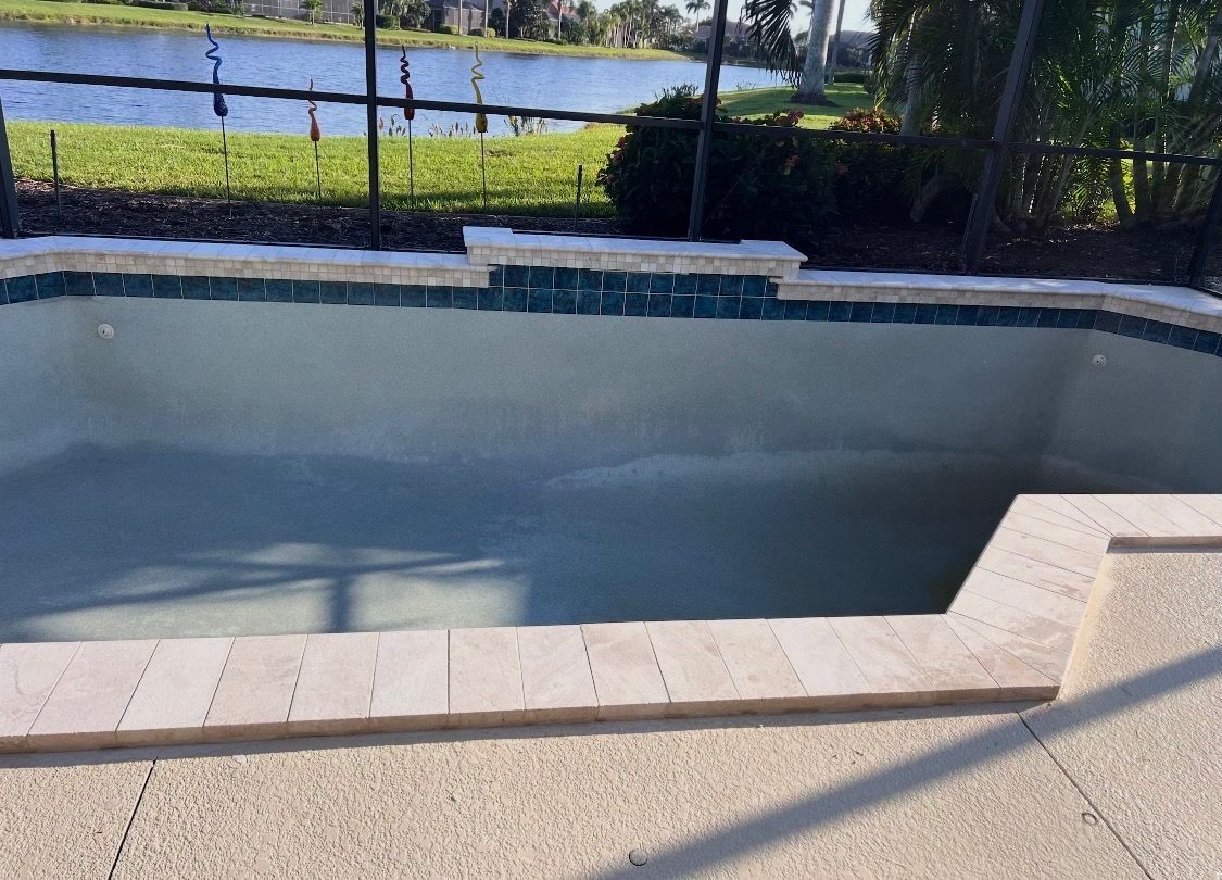 Empty swimming pool with light blue walls, stone deck, and sunlit water view beyond a fence