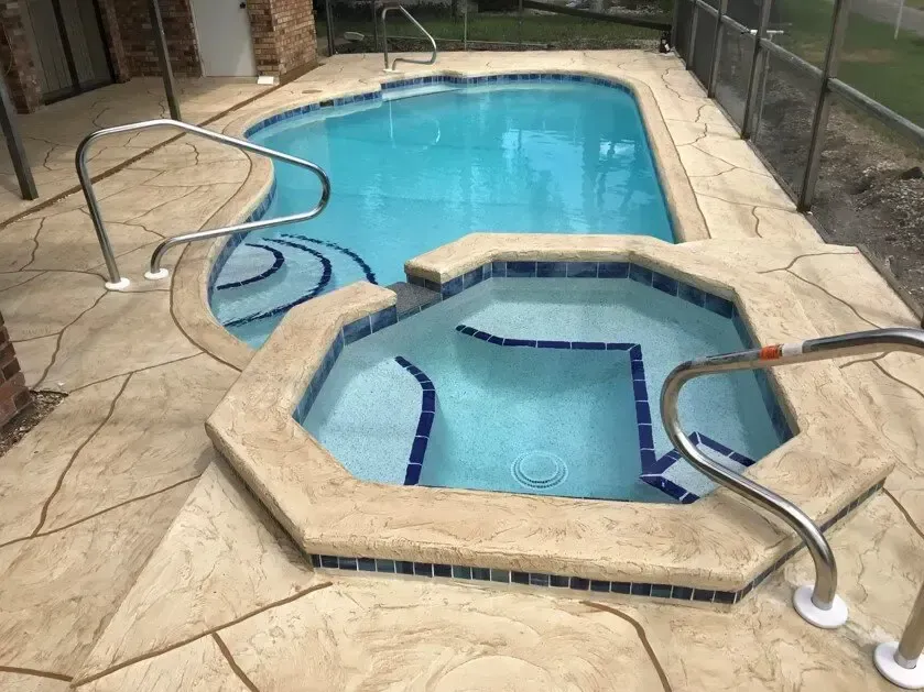 Pool and attached hot tub with blue water, tan stone deck, and metal handrails.