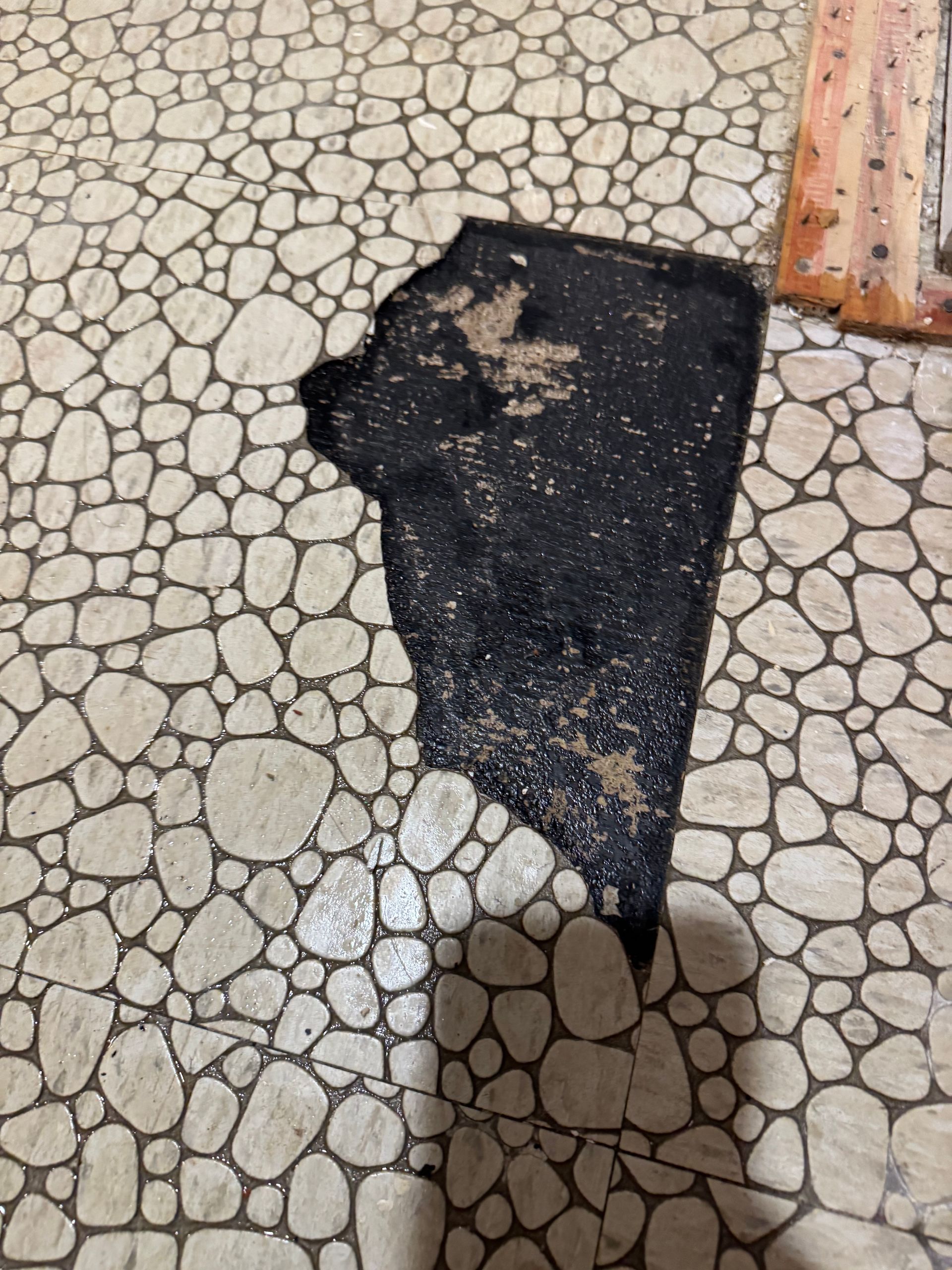 Hole in pebble tile flooring, revealing black underlayment. Adjacent to a textured, rectangular border.