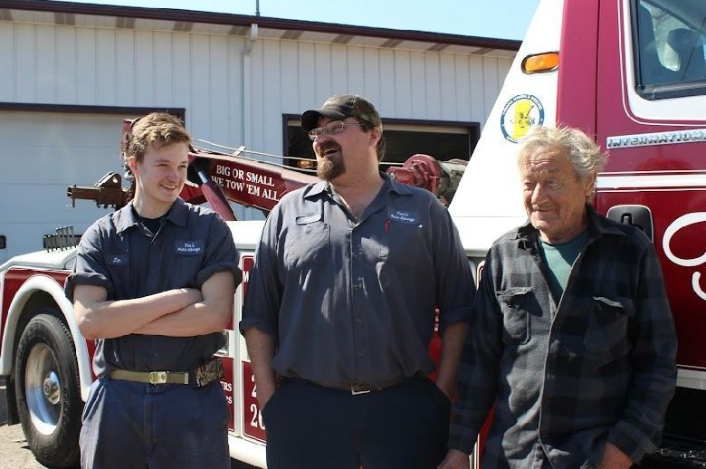 Three individuals stand smiling in front of a red tow truck parked outside a garage on a sunny day.