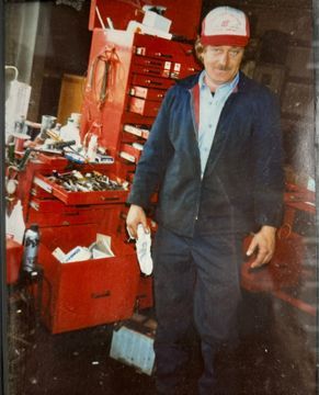 A person in a red cap and dark jacket stands in a garage beside a large red metal toolbox filled with various tools.