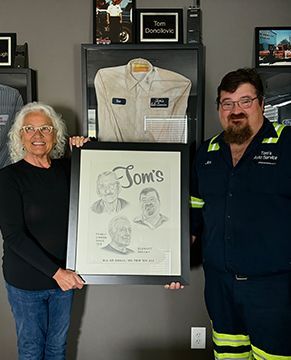 Two people holding a framed sketch of three faces in front of a display case containing a shirt, in an office setting.