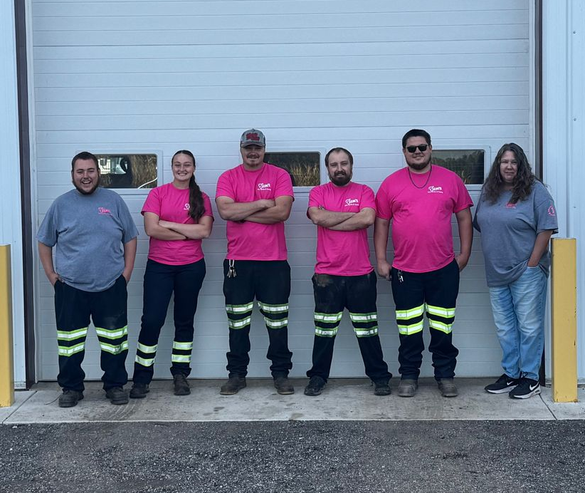 Six people in work attire stand in front of a garage door. Some wear pink shirts, others gray. Most have reflective stripes on pants.