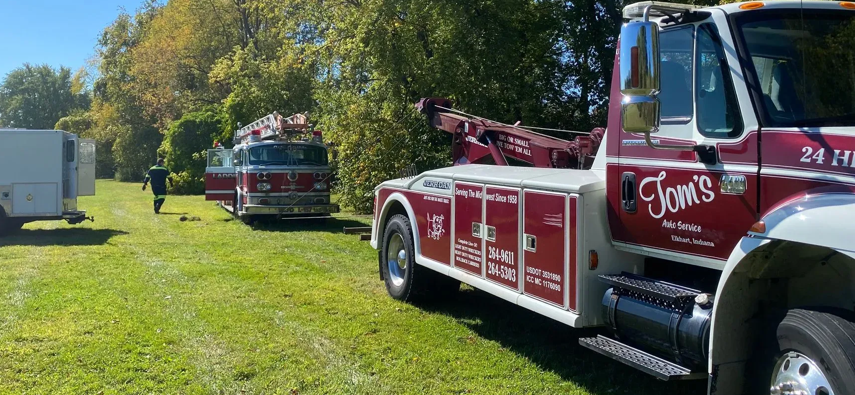 A red and white Tom’s tow truck parked on grass near a red fire truck and a white trailer on a sunny day.