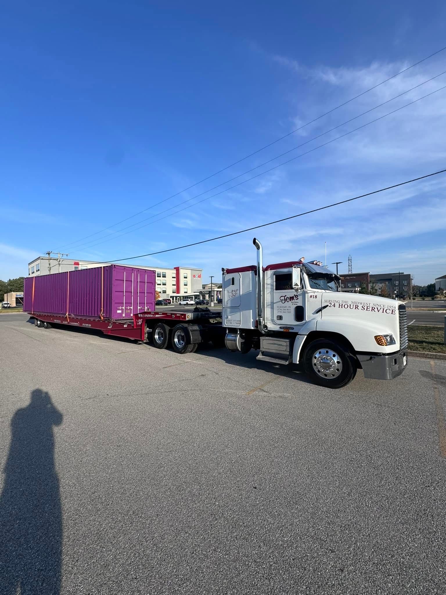 White semi-truck hauling large, purple cargo on a lowboy trailer; parked on gravel under a blue sky.